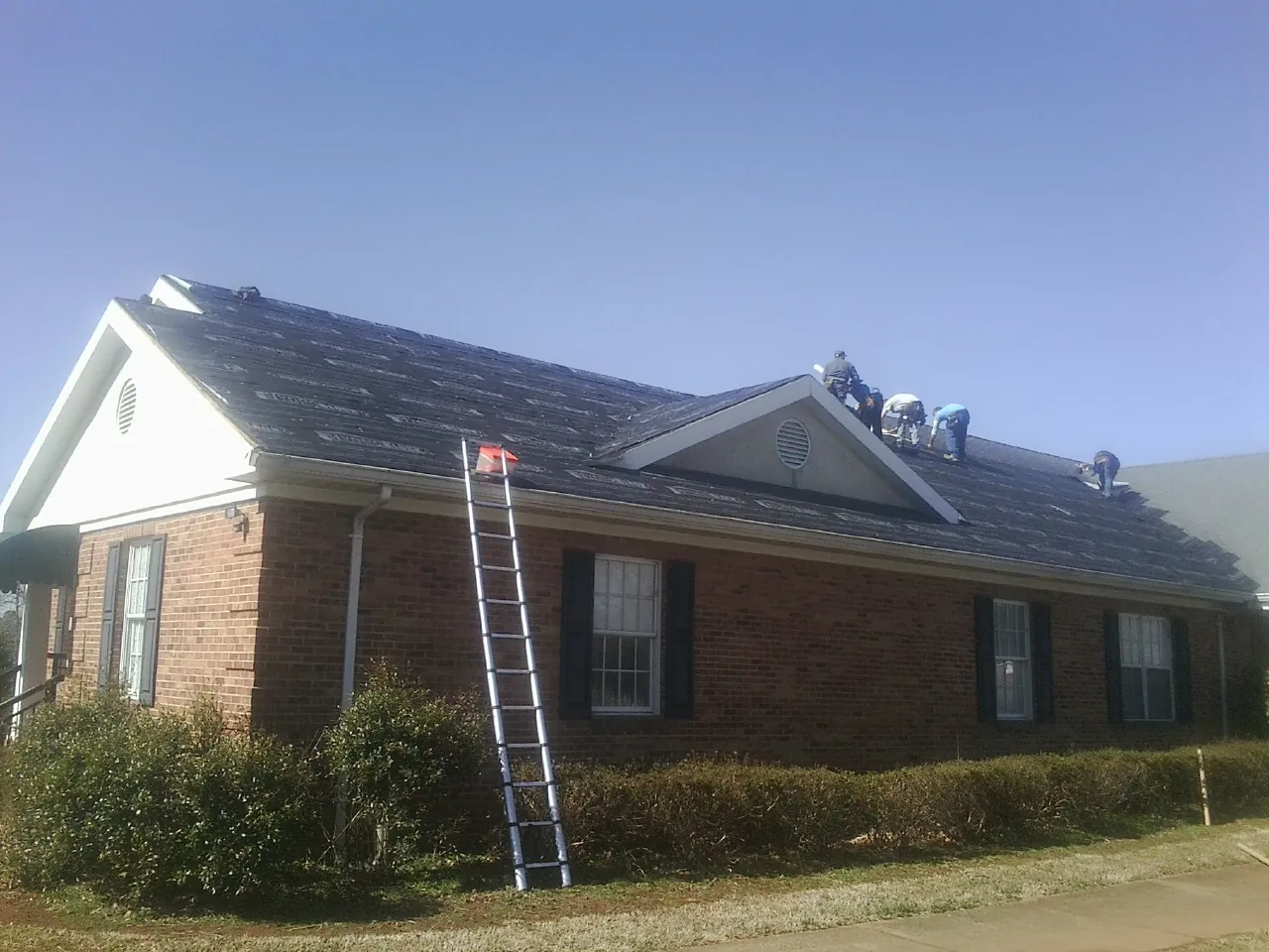 Crew tearing shingles off a brick ranch roof with a ladder set — Emergency Roof Repair work by our crew in Adena