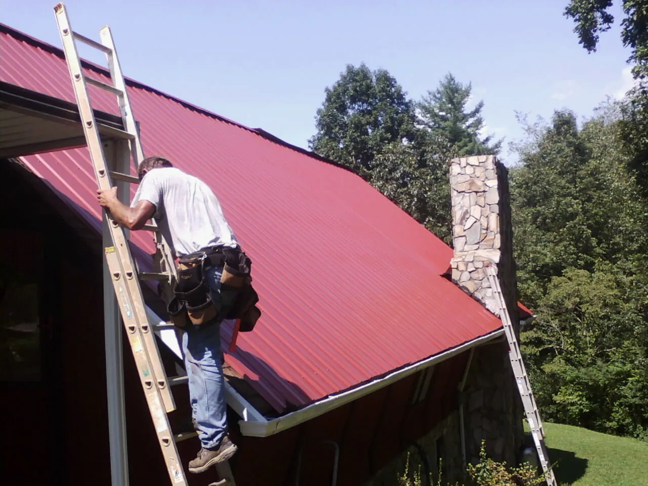 Roofer on a ladder beside a red metal roof and stone chimney — Soffit & Fascia Repair by Roofing Brothers in Adena